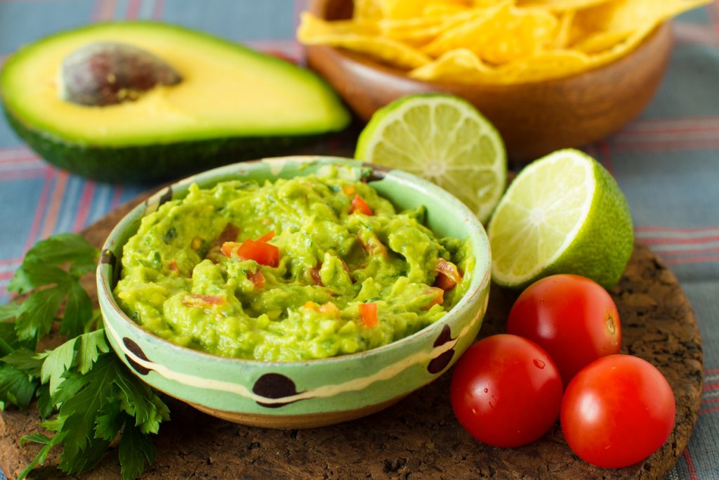 Schale mit cremiger Guacamole, umgeben von Limetten, Tomaten und einer Avocadohälfte.