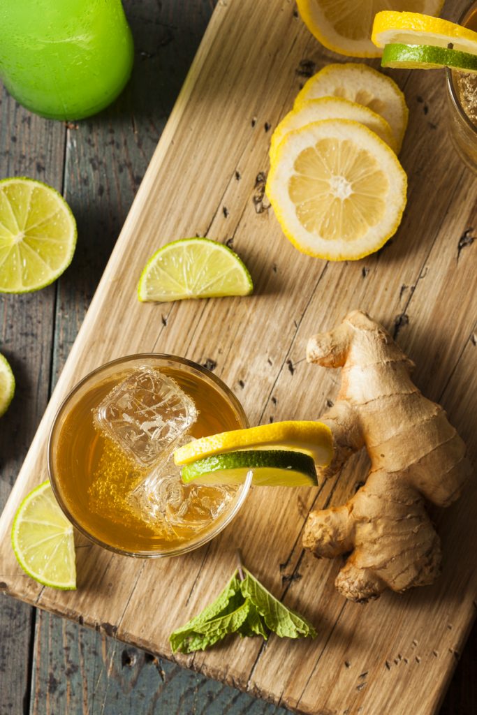 Glas Ginger Ale mit Eis, Zitronen- und Limettenscheiben sowie Ingwer auf Holzbrett.