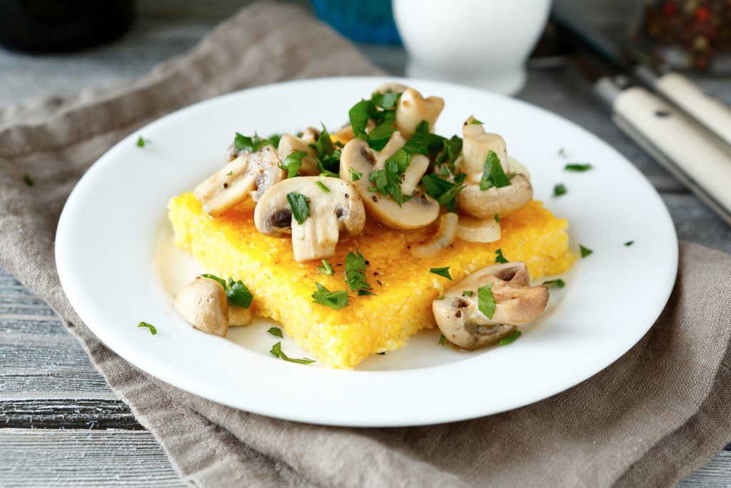 Gebratene Polentascheibe mit Zwiebeln, Champignons und Petersilie auf weißem Teller.