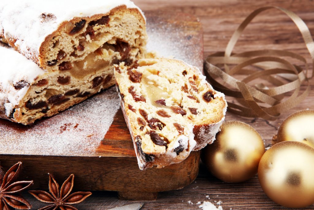 Ein angeschnittener Stollen mit Marzipanfüllung und Puderzucker liegt auf einem Holzbrett neben goldenen Kugeln.