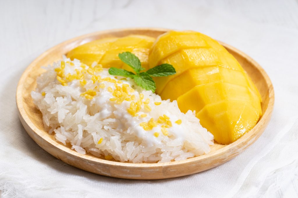 Mango Sticky Rice mit eingeschnittener Mango, Klebreis und Kokossauce auf einem runden Holzbrett.