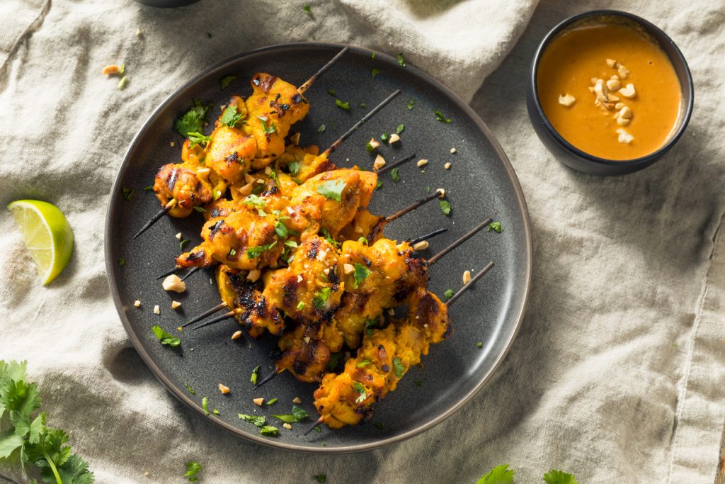 Teller mit mehreren Chicken Satay Spießen, daneben ein Schälchen Erdnusssauce, Limettenstück und Kräuter auf einem Tuch.