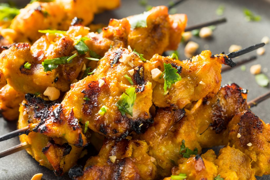 Nahaufnahme von gegrillten Chicken Satay Spießen mit goldbrauner Marinade, gehackten Erdnüssen und frischem Koriander auf einem Teller.