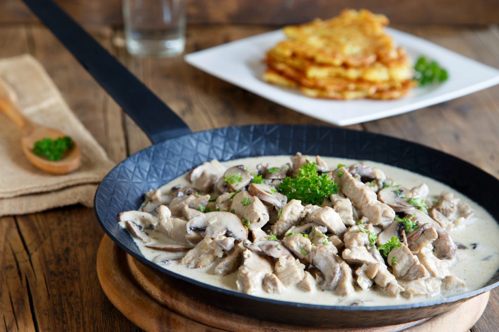 Züricher Kalbsragout mit Champignons liegt in einer Pfanne in heller Rahmsoße auf einem Holztisch.