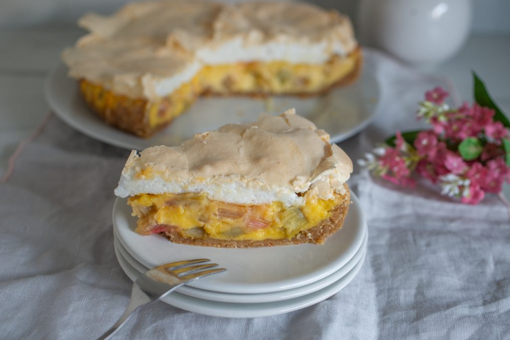 Ein Stück Kuchen mit heller Baiserhaube steht auf übereinander gestapelten Tellern vor der angeschnittenen Torte.