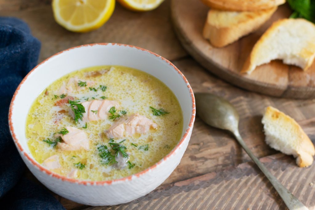 Schüssel mit Cremige Lachs-Sahnesuppe und Lachsstücken auf einem rustikalen Holztisch, daneben geröstetes Brot und Zitronenhälften.