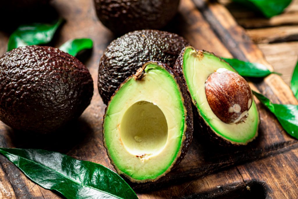 Halbierte und ganze Avocado liegen auf einem rustikalen Holzbrett zwischen grünen Blättern.