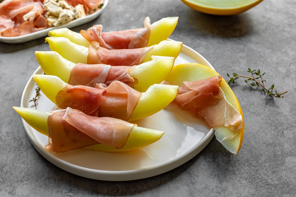 Auf einem Teller liegen mehrere Spalten Honigmelone, die jeweils mit dünnen Scheiben Prosciutto-Schinken umwickelt sind.