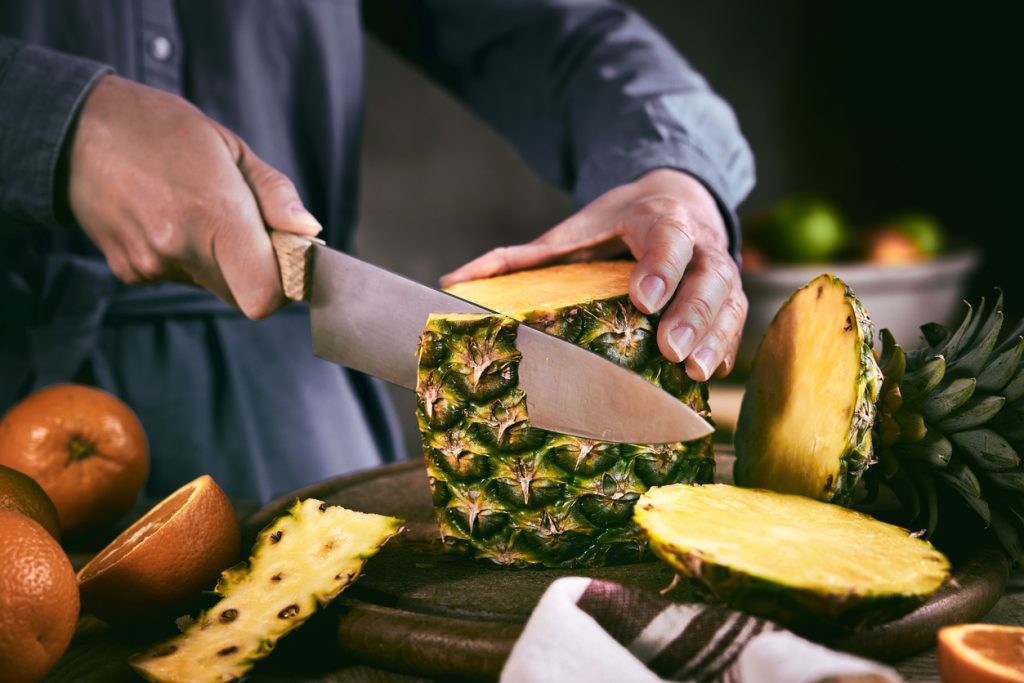 Eine Person schneidet mit einem großen Messer eine frische Ananas auf einem Holzbrett an.