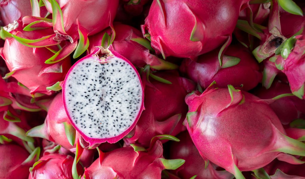 Viele pinke Pitahaya liegen dicht beieinander, eine Frucht ist halbiert und zeigt weißes Fruchtfleisch mit schwarzen Kernen.