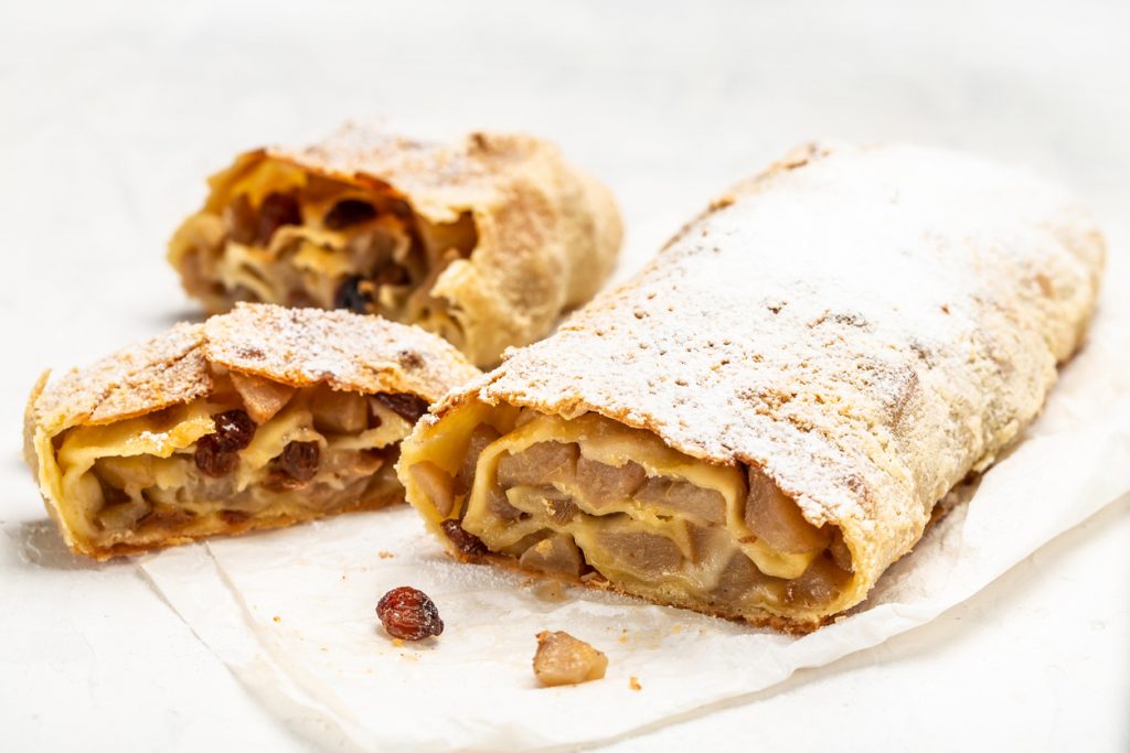 Ein langer hausgemachter Apfelstrudel liegt auf Backpapier und ist mit Puderzucker bestäubt, vorne sind angeschnittene Stücke zu sehen.