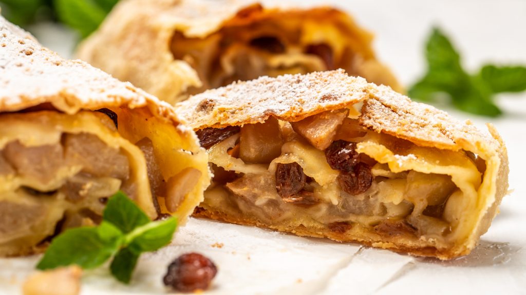 Eine aufgeschnittene Scheibe hausgemachter Apfelstrudel liegt vorne im Fokus und zeigt eine saftige Füllung aus Apfelstücken und Rosinen.