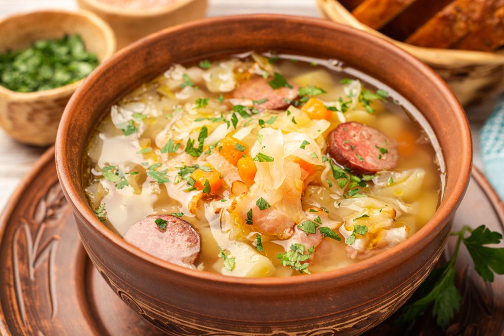 Polnische Sauerkrautsuppe Kapusniak mit Kartoffeln, Sauerkraut und Wurstscheiben in einer braunen Schüssel.