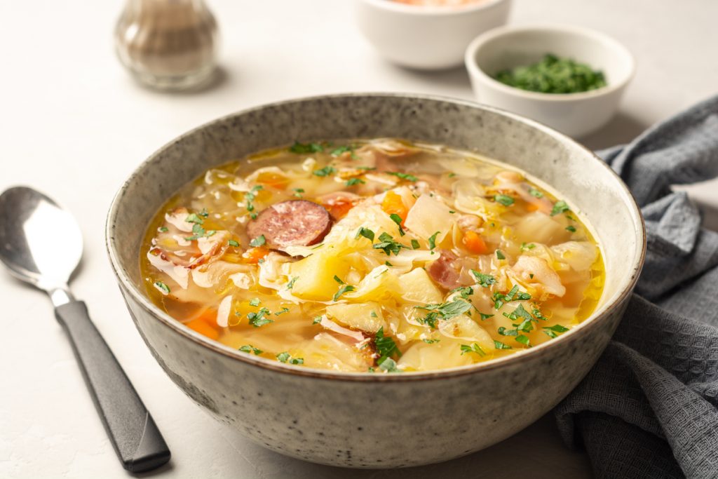 Eine Schale Polnische Sauerkrautsuppe Kapusniak steht angerichtet mit Besteck und Kräuterschälchen im Hintergrund.
