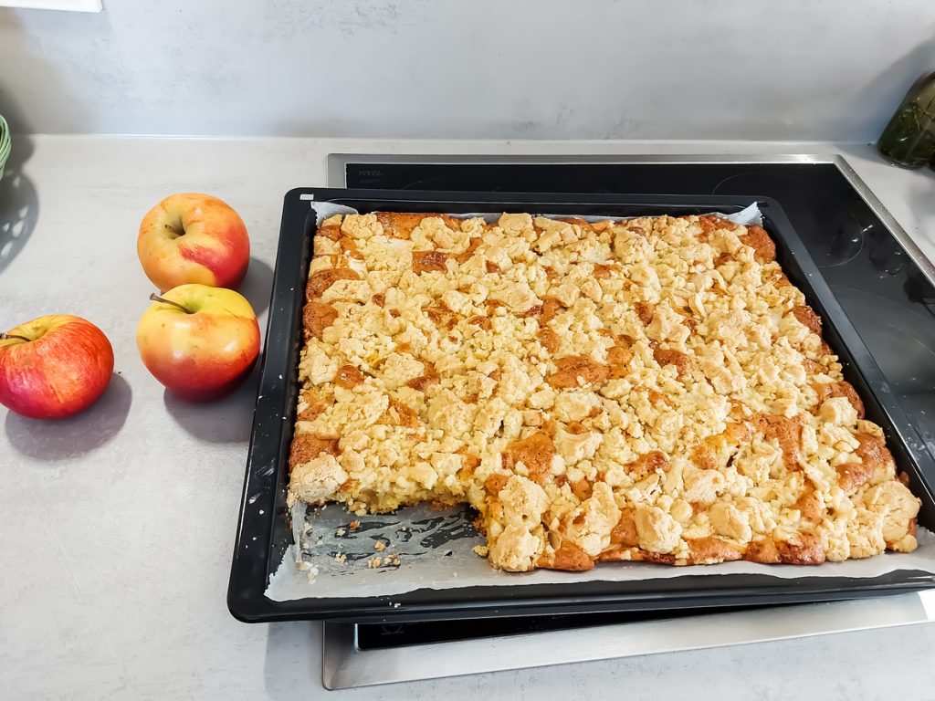 Ein großes Blech Apfelstreuselkuchen vom Blech steht auf dem Herd, daneben liegen drei frische Äpfel.