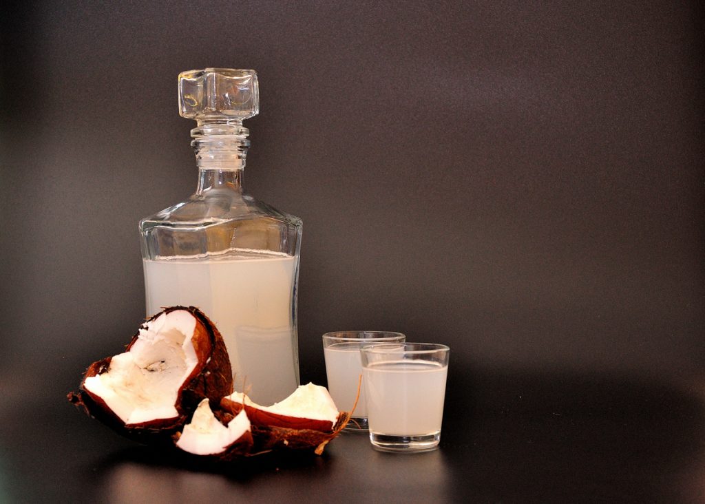 Milchig-weißer Kokoslikör steht in einer Glasflasche neben zwei kleinen Gläsern und Kokosstücken auf dunklem Hintergrund.