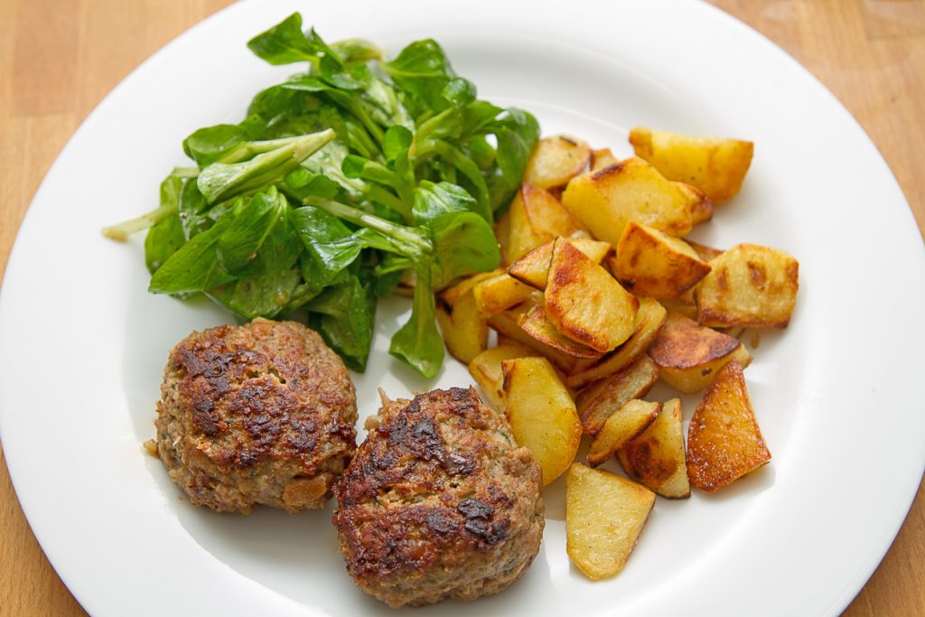 Auf einem weißen Teller liegen zwei knusprige Bouletten mit goldbraunen Bratkartoffeln und frischem Feldsalat.