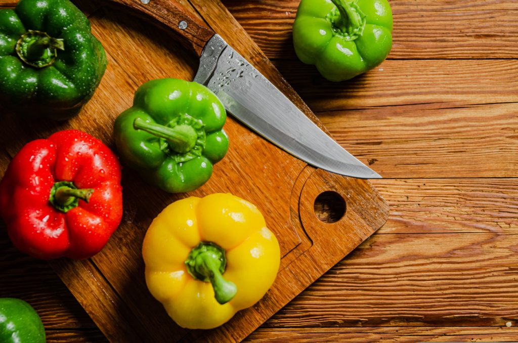 Mehrere grüne, gelbe und rote Paprika liegen mit einem Messer auf einem Holzbrett.