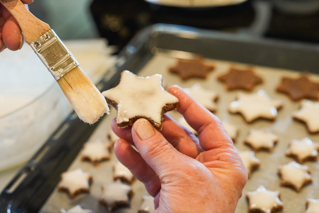 Eine Hand hält einen Zimtstern und bestreicht ihn mit weißer Glasur, im Hintergrund liegt ein Blech voller Sterne.
