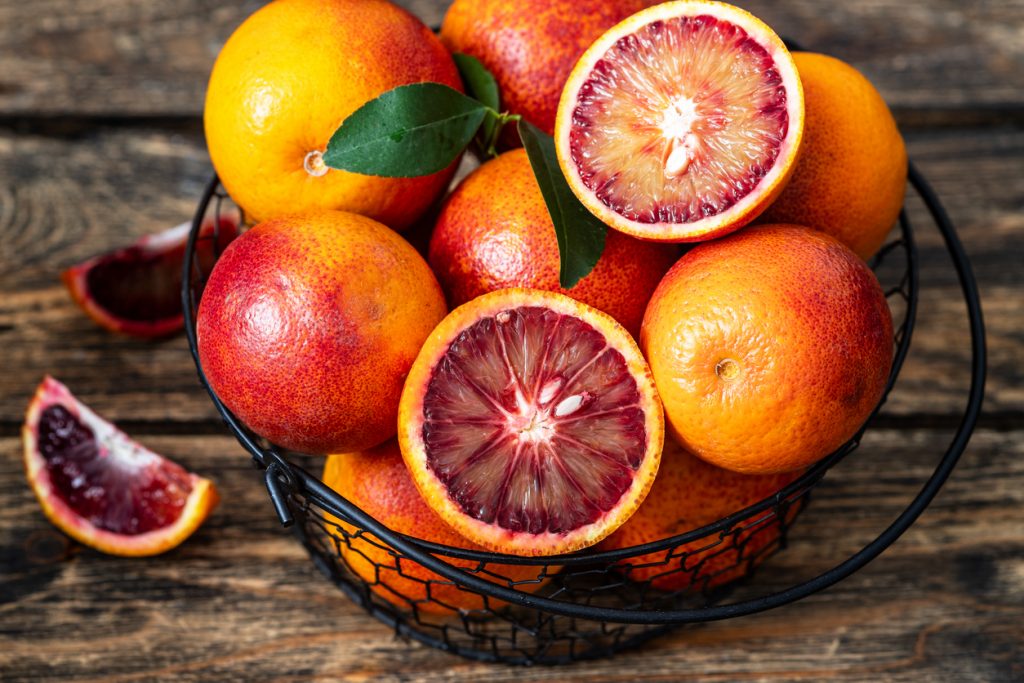 Ein schwarzer Drahtkorb ist mit ganzen und aufgeschnittenen Blutorangen gefüllt, die auf einem Holztisch liegen.