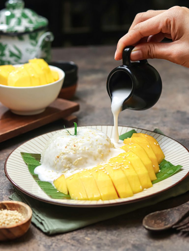 Eine Hand gießt Kokossauce über Mango Sticky Rice mit Klebreis und Mangoscheiben.