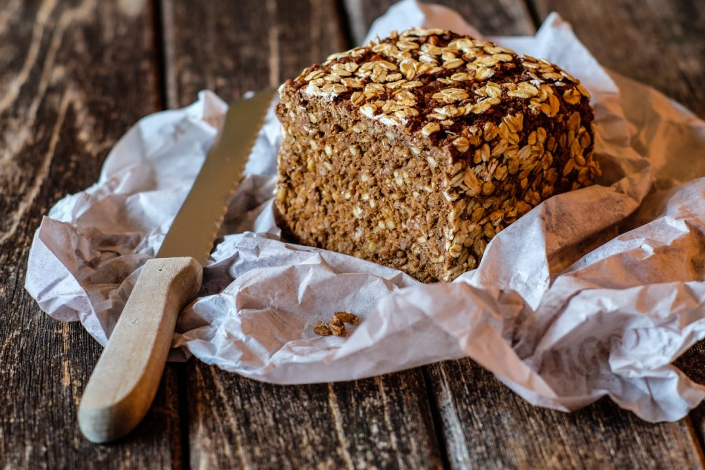 Ein angeschnittenes Roggenvollkornbrot mit Haferflockenkruste liegt auf Backpapier neben einem Brotmesser.