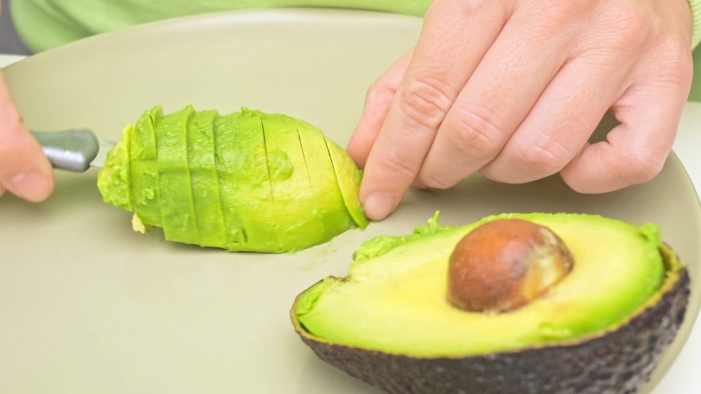 Hände schneiden eine reife Avocado auf einem Schneidebrett in Stücke.