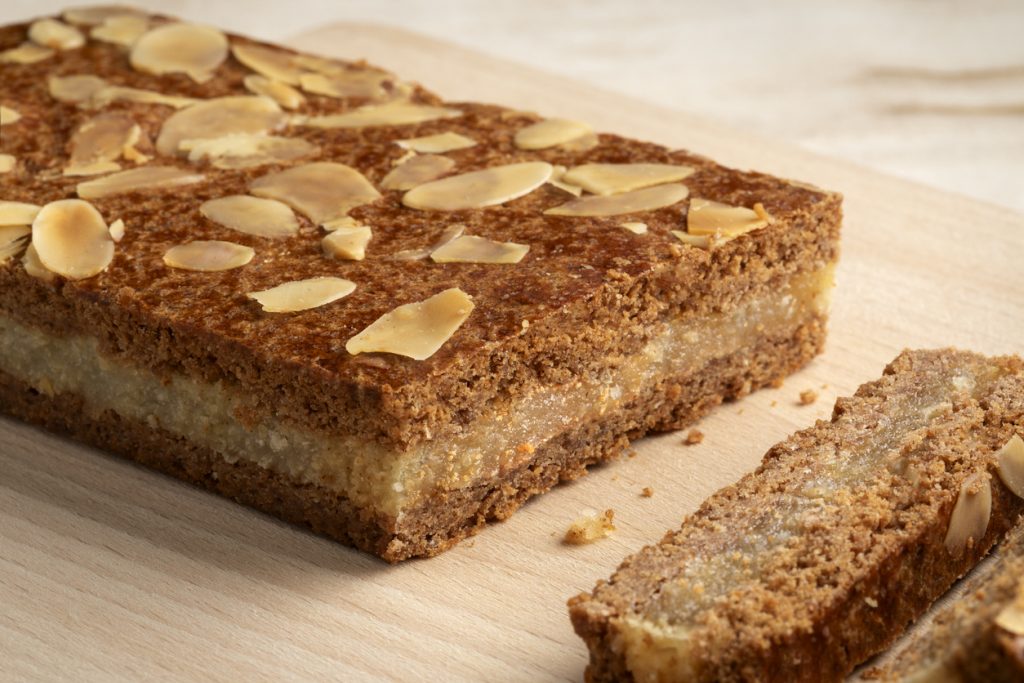 Ein länglicher Gevulde Spekulatius mit heller Füllung und Mandelblättchen liegt auf einem Holzbrett, ein Stück ist bereits angeschnitten.