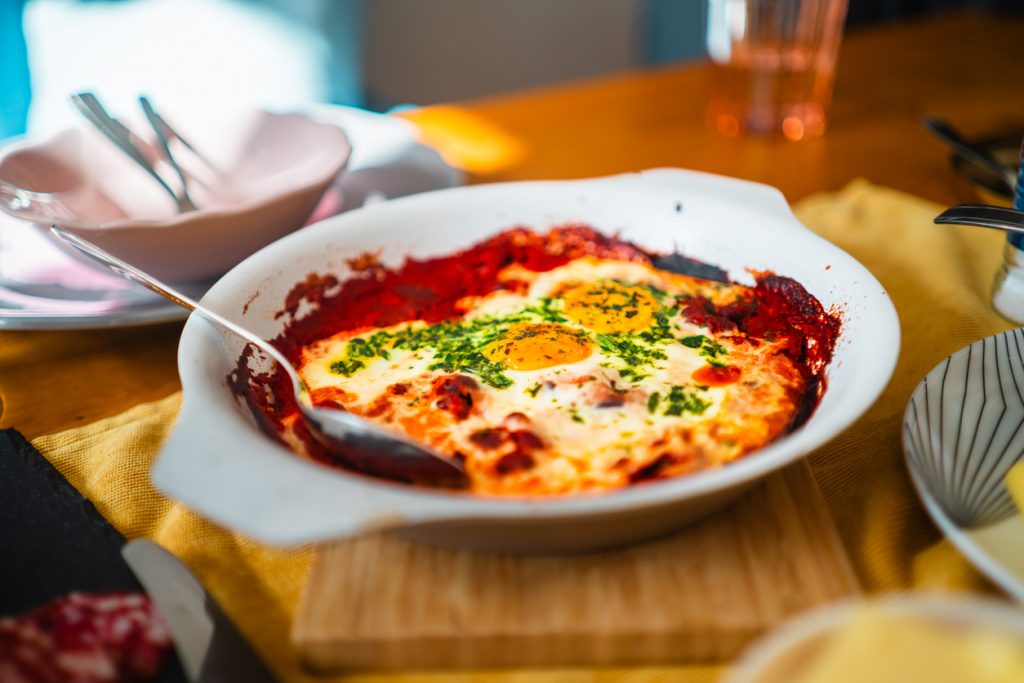 Weiße Auflaufform mit Eiern in tomatiger Sauce auf einem gedeckten Tisch.