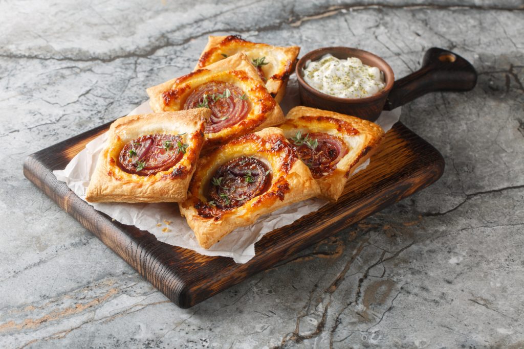 Ein Holzbrett ist mit vier Blätterteigtaschen mit karamellisierter Zwiebel und einem kleinen Schälchen Dip angerichtet.