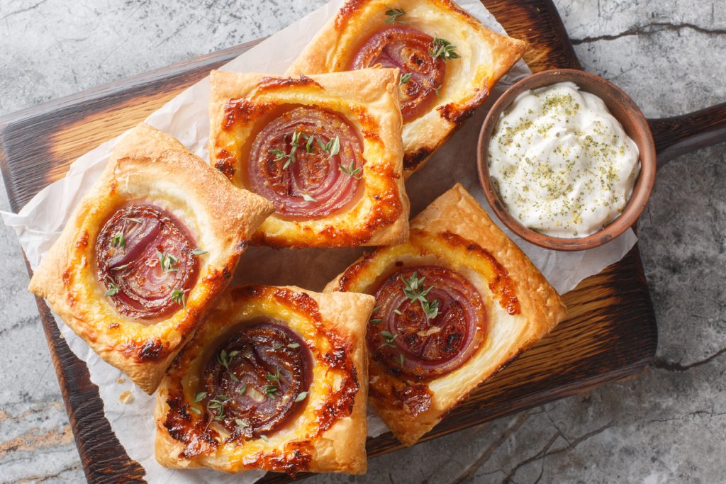 Mehrere quadratische Blätterteigtaschen mit karamellisierter Zwiebel und Käse liegen auf einem Holzbrett neben einem Schälchen cremigem Dip.