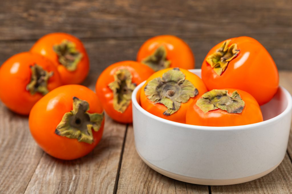 Mehrere Kaki liegen in einer weißen Schale auf einem Holztisch.