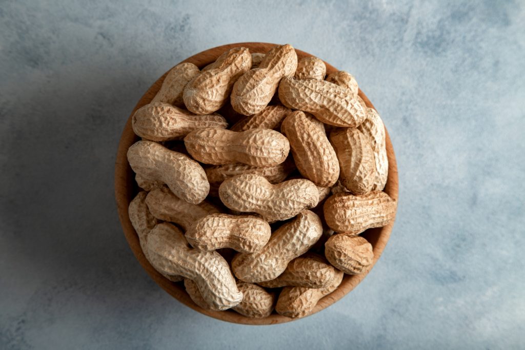 Holzschale von oben fotografiert, gefüllt mit ungeschälten Erdnüssen auf hellblauem Untergrund.