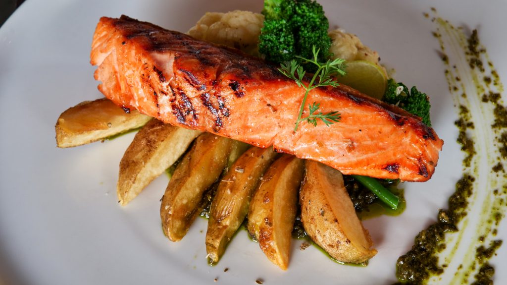 Ein Stück gegrillter Lachs liegt auf Bratkartoffeln mit grünem Gemüse und einer Spur Kräutersauce auf einem weißen Teller.