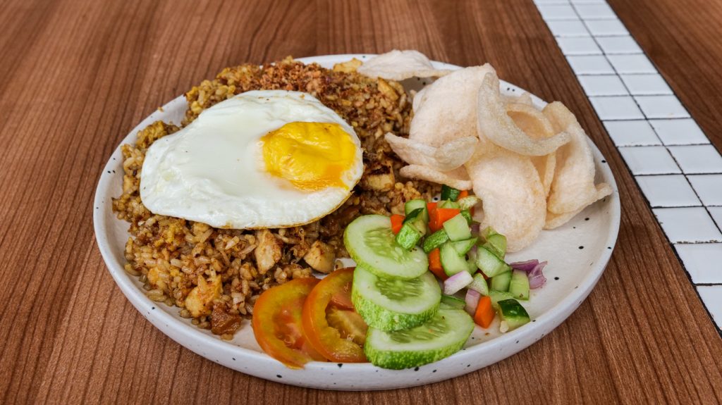 Indonesischer Nasi Goreng mit Spiegelei, Gurken-Tomaten-Salat und vielen Krabbenchips auf einem hellen Teller.