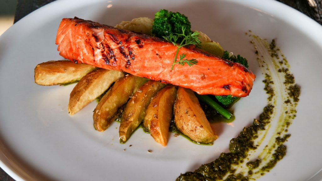 Ein längliches Lachsfilet liegt auf knusprigen Kartoffelspalten mit Brokkoli, Gemüse und Kräutersauce auf einem großen weißen Teller.