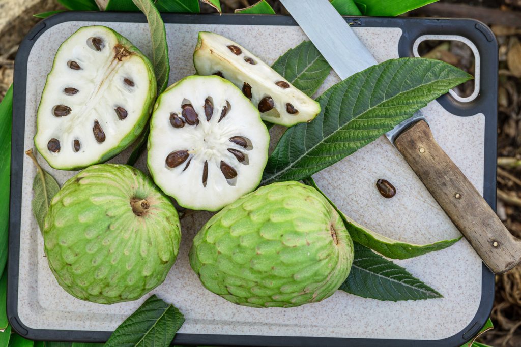 Eine Cherimoya ist halbiert und liegt mit Kernen neben einem Messer auf einem Brett.