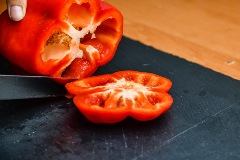 Eine rote Paprika wird auf einem dunklen Brett mit einem Messer in Scheiben geschnitten.