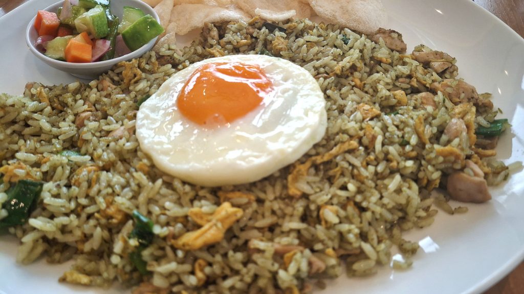 Teller mit Nasi Goreng, darauf ein Spiegelei, dazu Gurken-Gemüsewürfel und Krabbenchips.