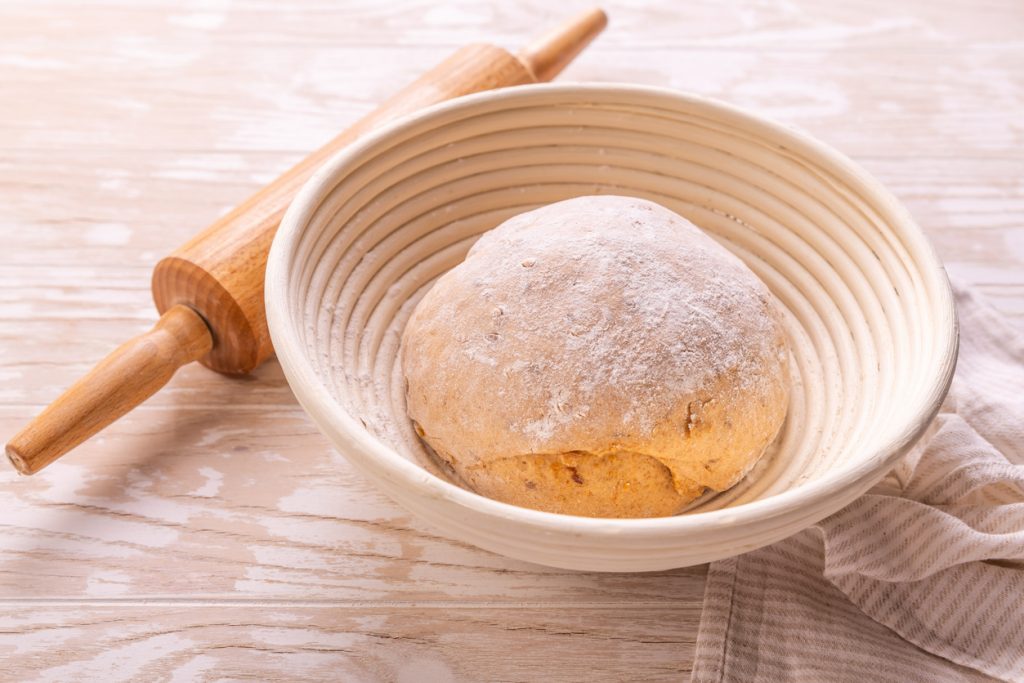Ein bemehlter Hefeteigball liegt in einem runden Gärkörbchen, daneben liegt ein Holz-Nudelholz.