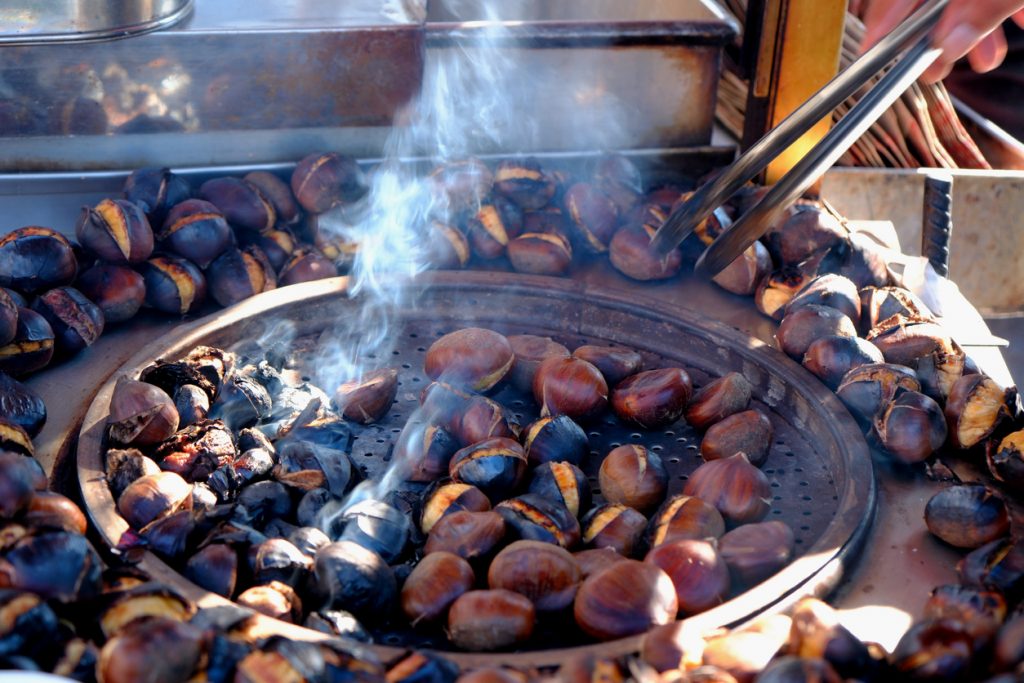 Maronen rösten in einer großen Pfanne, während Rauch aufsteigt und eine Zange sie bewegt.