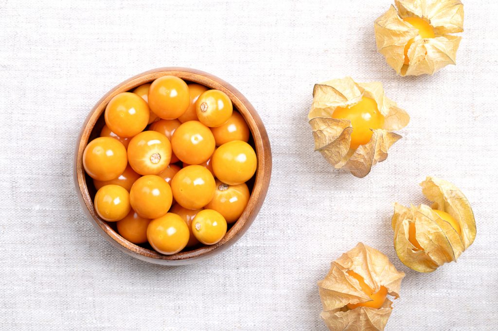 Holzschale mit vielen orangefarbenen Beeren neben geöffneten Hüllen auf hellem Stoff.