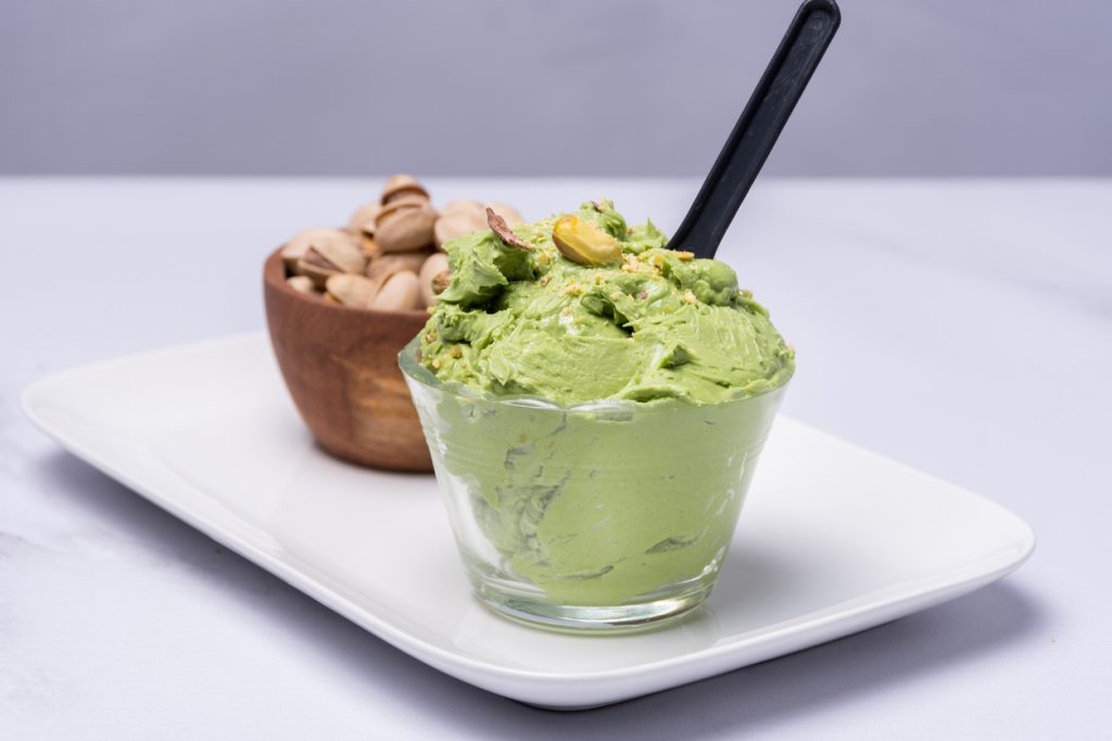 Pistazieneis in einem Glas mit Löffel und Pistazien im Hintergrund.