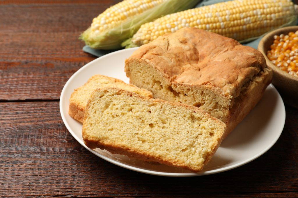Maisbrot liegt als Laib mit angeschnittenen Scheiben auf einem weißen Teller, daneben sieht man Maiskolben.