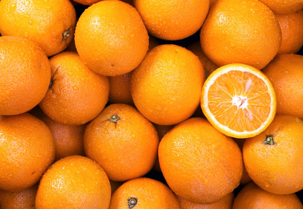 Viele ganze Orangen mit Wassertropfen liegen dicht beieinander, eine Orange ist halbiert und zeigt das Fruchtfleisch.