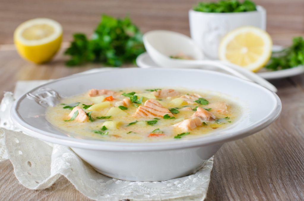 Tiefer weißer Teller mit Cremige Lachs-Sahnesuppe, in der Lachswürfel, Kartoffelstücke und Petersilie schwimmen, im Hintergrund Zitronenhälften und Kräuter.