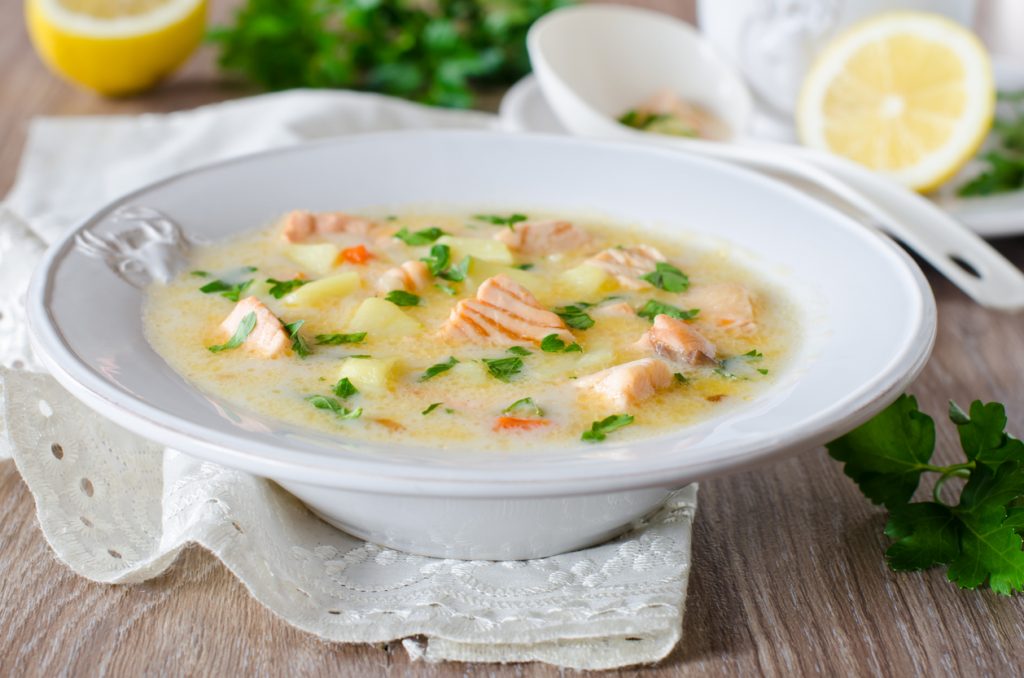 Weißer Teller mit Cremige Lachs-Sahnesuppe, darin Lachswürfel, Kartoffeln und Kräuter, im Hintergrund Zitronen und ein Kräutertopf.