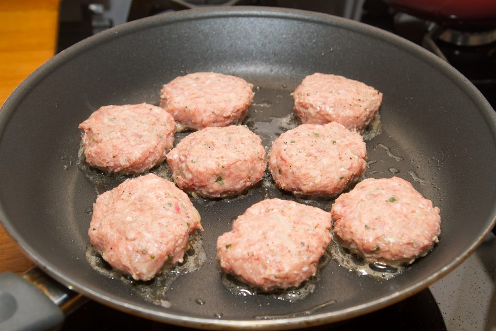 Acht noch rohe Bouletten liegen in einer Pfanne und werden gerade im heißen Fett angebraten.