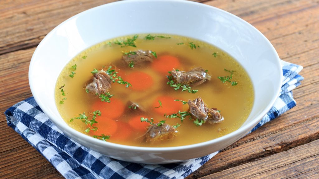 Tiefer weißer Teller mit klarer Rinderbrühe, Rindfleischstückchen, Karottenscheiben und Petersilie auf einem Holztisch.