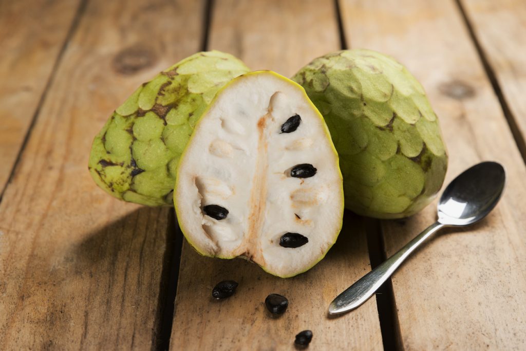 Eine halbierte Cherimoya liegt auf einem Holztisch neben einem Löffel und einzelnen Kernen.